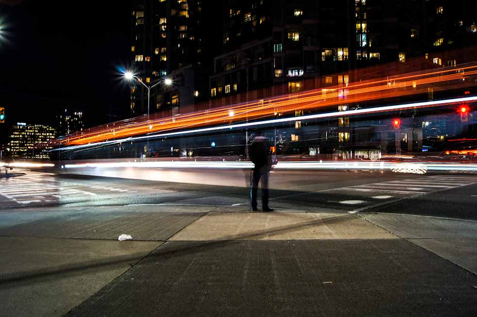 Person standing on a busy street corner with long-exposure car lights whizzing by